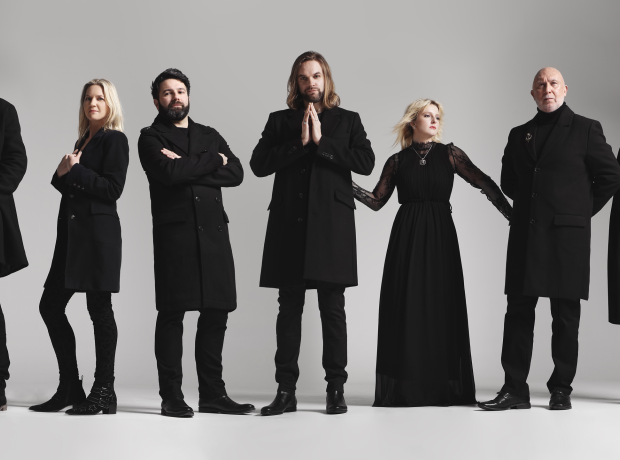 De band Rumours of Fleetwood Mac poseert voor een lichte achtergrond. Zeven leden in zwarte kleding staan naast elkaar, met variërende houdingen die een stijlvolle en dramatische uitstraling geven.