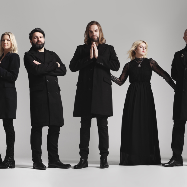 De band Rumours of Fleetwood Mac poseert voor een lichte achtergrond. Zeven leden in zwarte kleding staan naast elkaar, met variërende houdingen die een stijlvolle en dramatische uitstraling geven.