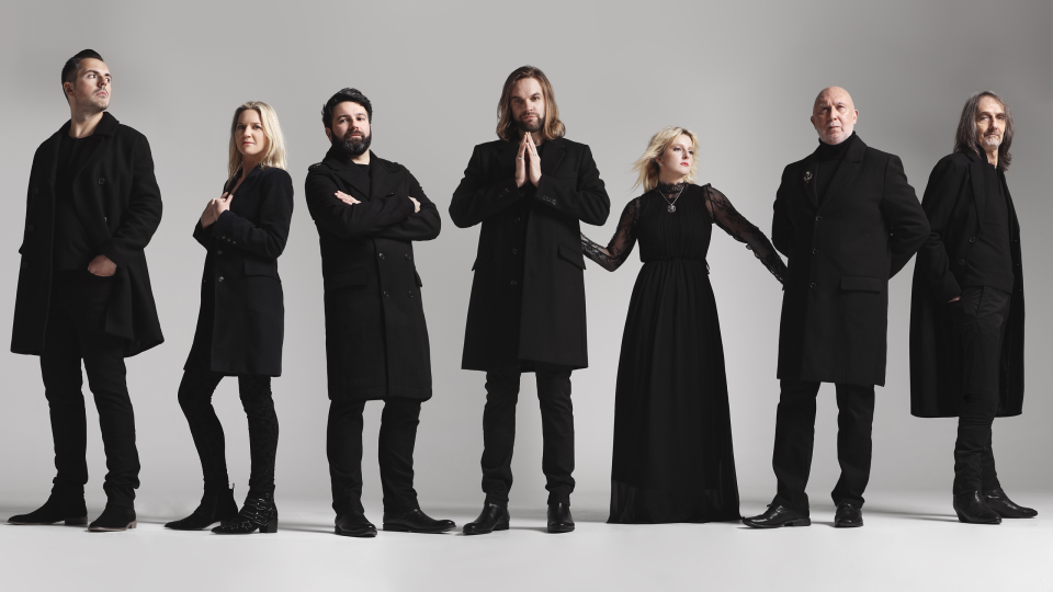 De band Rumours of Fleetwood Mac poseert voor een lichte achtergrond. Zeven leden in zwarte kleding staan naast elkaar, met variërende houdingen die een stijlvolle en dramatische uitstraling geven.